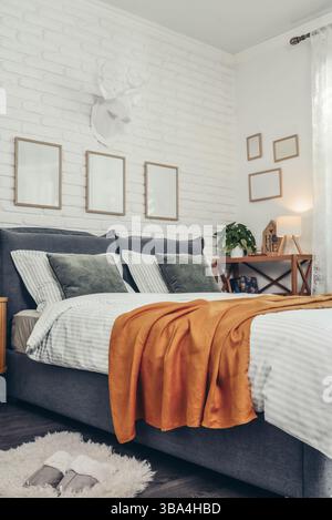 Interni eleganti della camera da letto con grande e comodo letto grigio, tovaglia di senape e comodino in legno vicino alle pareti in mattoni bianchi Foto Stock