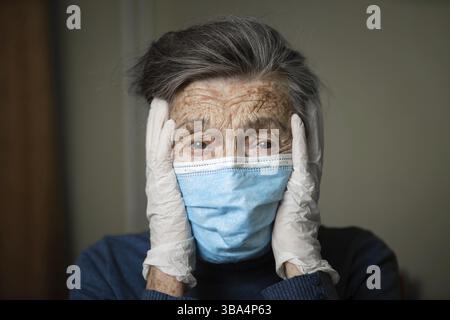 Ritratto di una donna caucasica anziana con rughe, indossando una maschera medica e guanti, l'emozione della fragilità e imprevedibilità dovuta al coronav Foto Stock