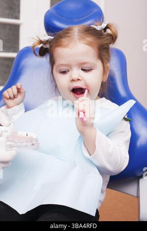 Due anni ragazza è imparare a lavarsi i denti con spazzolino da denti in mano in studio dentistico Foto Stock