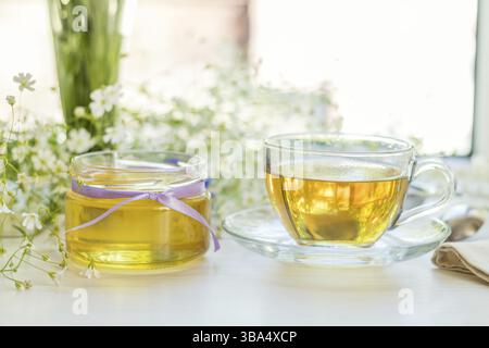Miele in vasetti di vetro e tazza di tè con fiori bianchi di primavera su davanzale. Profondità di campo ridotta Foto Stock