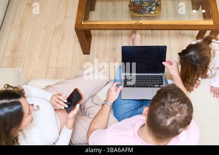 Padre che mostra qualcosa al laptop a sua figlia mentre la madre incinta usa il suo smartphone, la famiglia che passa il tempo insieme a casa Foto Stock