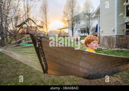 Il ragazzo siede felicemente sull'amaca al tramonto il giorno di primavera nel cortile Foto Stock