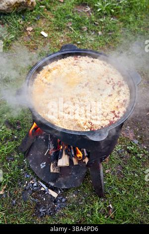 Vista dall'alto del processo di cottura del pilaf su un fuoco aperto in un calderone su una griglia, con riso bollente e vapore crescente, all'aperto in estate Foto Stock