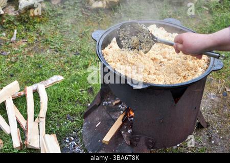Vista dall'alto del processo di cottura del pilaf su un fuoco aperto in un calderone su una griglia, con riso bollente e vapore crescente, all'aperto in estate Foto Stock