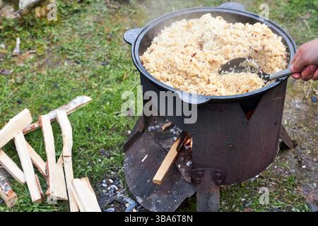 Vista dall'alto del processo di cottura del pilaf su un fuoco aperto in un calderone su una griglia, con riso bollente e vapore crescente, all'aperto in estate Foto Stock
