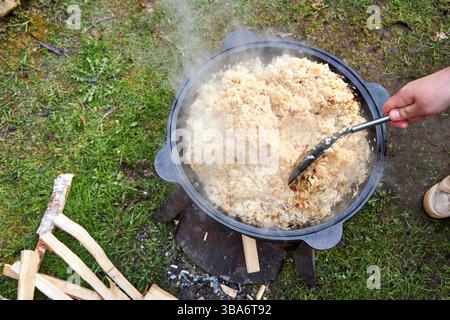 Vista dall'alto del processo di cottura del pilaf su un fuoco aperto in un calderone su una griglia, con riso bollente e vapore crescente, all'aperto in estate Foto Stock