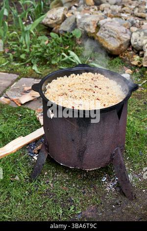 Vista dall'alto del processo di cottura del pilaf su un fuoco aperto in un calderone su una griglia, con riso bollente e vapore crescente, all'aperto in estate Foto Stock