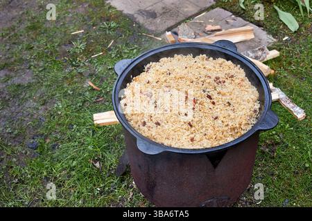 Vista dall'alto del processo di cottura del pilaf su un fuoco aperto in un calderone su una griglia, con riso bollente e vapore crescente, all'aperto in estate Foto Stock