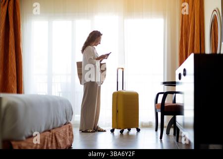 Donna in camera d'albergo con una valigia gialla, che guarda il suo telefono Foto Stock