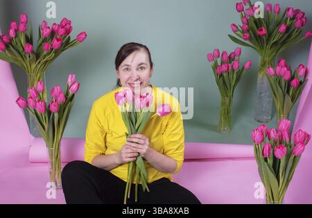 Una donna di mezza età felice con un mucchio di tulipani è felice e sorridente su uno sfondo rosa-verde Foto Stock