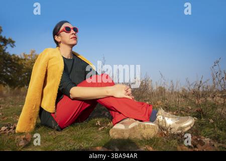 Una donna di mezza età vestita alla moda siede nell'erba contro un cielo blu Foto Stock