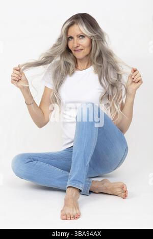 Bella donna adulta con lunghi capelli grigi che indossa una T-shirt bianca e jeans su sfondo bianco Foto Stock