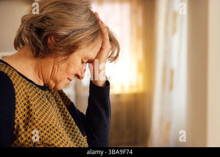 Primo piano di una donna anziana che soffre di mal di testa. La signora matura ha chiuso gli occhi e tiene la fronte con la mano. Un pensionato in casua Foto Stock