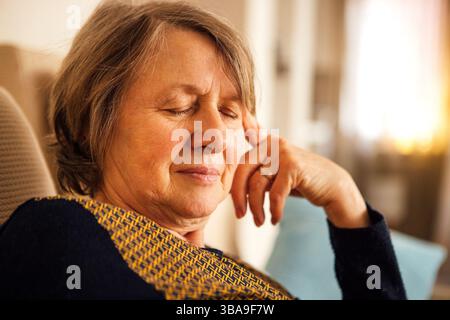 Primo piano di una donna anziana che soffre di mal di testa. La signora matura ha chiuso gli occhi e tiene la fronte con la mano. Un pensionato in casua Foto Stock
