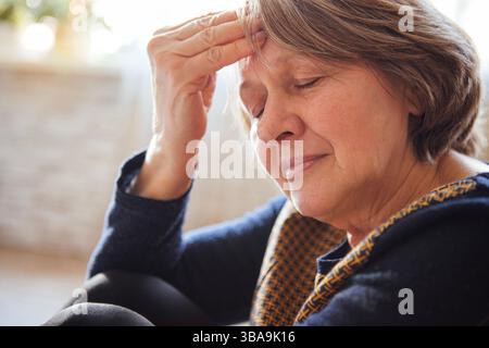 Primo piano di una donna anziana che soffre di mal di testa. La signora matura ha chiuso gli occhi e tiene la fronte con la mano. Un pensionato in casua Foto Stock
