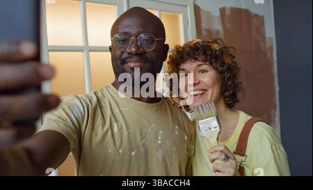 Coppia felice che scatta selfie mentre dipinge una stanza, sorride e si diverte a un progetto di ristrutturazione della casa, entrambi con strumenti di pittura, sembrano entusiasti e orgogliosi del progresso Foto Stock