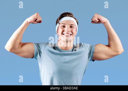 Positivo giovane sportivo seduto su palla fitness e mostrandogli forza su sfondo lilla studio Foto Stock