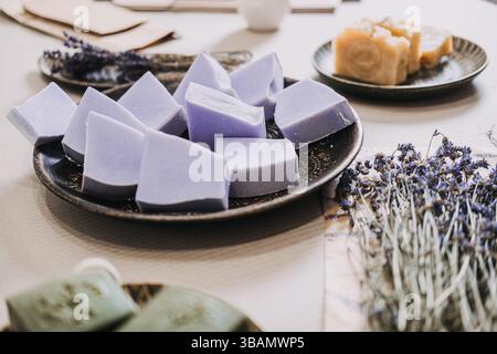 Primo piano di saponette fatte a mano disposte su un piatto di ceramica con lavanda secca e sapone verde nelle vicinanze. Aromaterapia, bellezza pulita, sostenibile Foto Stock