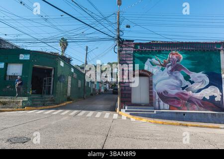 Uomo in un angolo di strada vicino a una casa con murale di una signora con la veste bianca vorticosa a San Marcos de Colon, dipartimento di Chaluteca, Honduras Foto Stock