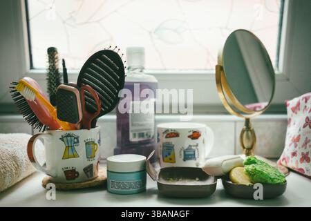 Articoli per l'igiene personale sul ripiano in bagno, pettini e spazzole in una tazza, creme, saponi e uno specchio. Foto Stock
