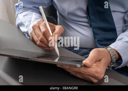 Primo piano di mani maschili mature utilizzando uno stilo bianco su un tablet, che illustra la firma elettronica, il flusso di lavoro digitale e la moderna tecnologia aziendale. Foto Stock