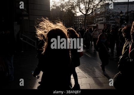 Persone che camminano attraverso una strada della città di Barcellona, in Spagna, in una giornata molto ventosa, con raffiche forti, silhouette retroilluminate e luce del sole pomeridiano. Foto Stock