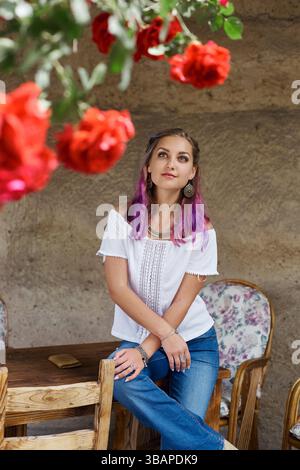 una giovane donna si appoggia su un tavolo di legno, indossando un top bianco e jeans blu. Ammira le vivaci rose rosse appese sopra in un ambiente rustico pieno Foto Stock