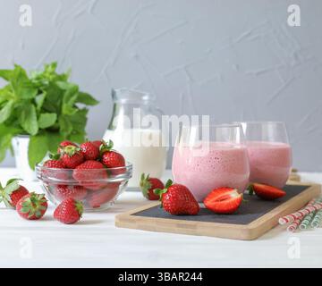Due bicchieri di frullato di fragole posizionati sul piatto da portata accanto alla ciotola di fragole fresche e una caraffa di latte, circondati da foglie di menta, creando un'atmosfera vivace. Foto Stock