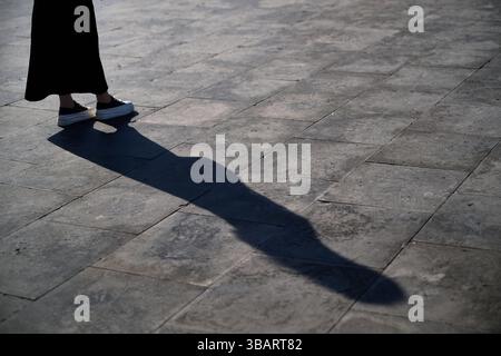 La lunga ombra di una donna si estende sul marciapiede di pietra, poiché solo le sue scarpe e la sua gonna sono visibili, evocando mistero. Foto Stock