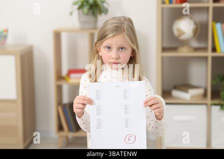 Grado scolastico. Ragazza triste che tiene in mano il foglio delle risposte con la lettera F in casa Foto Stock