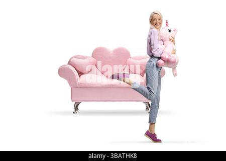 Donna felice che tiene in mano un grande unicorno giocattolo davanti a un divano rosa isolato su sfondo bianco Foto Stock