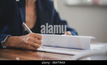 Donna in un blazer che esamina documenti finanziari, contratti e grafici, prende appunti durante una riunione o una sessione di lavoro. Foto Stock