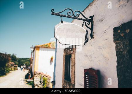 Un modello di tabellone vintage vuoto appeso a una staffa di ferro ornata su un vecchio muro di pietra imbiancato in un pittoresco villaggio illuminato dal sole, perfetto per un marchio Foto Stock