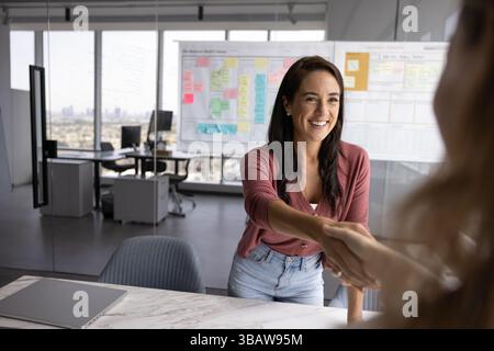 Giovane donna d'affari amichevole che stringe la mano con una compagna sul tavolo Foto Stock