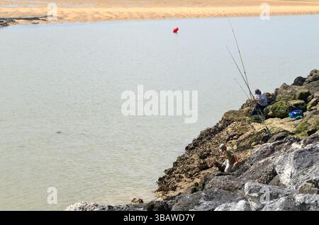 La Palmyre, Francia - 1° maggio 2025: Due pescatori pescano dalle rocce in riva all'acqua, circondati da un tranquillo paesaggio di spiaggia e da dolci onde Foto Stock