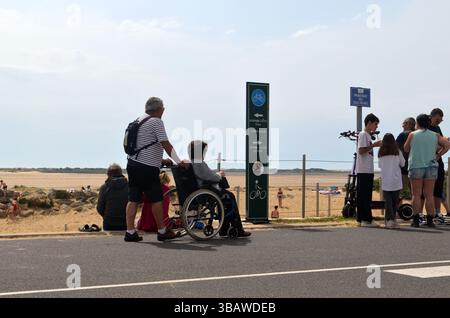 La Palmyre, Francia - 1° maggio 2025: Le persone si radunano in spiaggia, con una sedia a rotelle in primo piano che osserva la vibrante atmosfera estiva Foto Stock