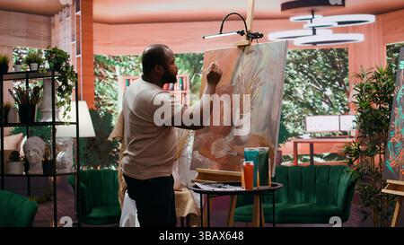 Artista afroamericano che usa la pittura ad acquerello e mescola i colori su tavolozza per la sua creazione artistica in un laboratorio di belle arti. Uomo che dipinge elementi rustici su tela, processo creativo. Telecamera B.. Foto Stock