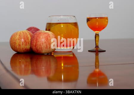 primo piano di tre mele fresche appoggiate su un tavolo di legno lucido, accompagnate da due bicchieri pieni di succo di mela dorato. Il rosso vibrante e urla Foto Stock