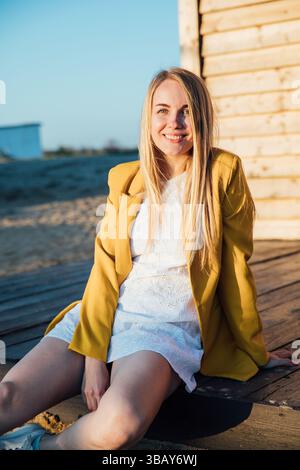 Donna bionda in abito bianco e giacca gialla seduta vicino a una casa di legno Foto Stock
