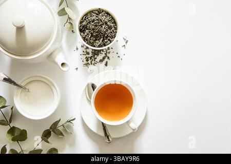 Dal tè appena preparato fuoriesce vapore delicato in tazza bianca accanto alle foglie di erbe nel recipiente e allo zucchero nel piatto, circondato da rami di eucalipto su sfondo bianco brillante. Foto Stock