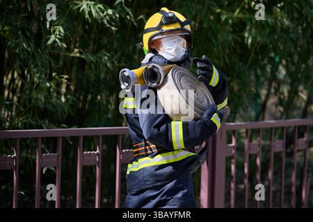 Vigile del fuoco che trasporta un tubo a spirale durante un'esercitazione alla luce del giorno, indossando l'attrezzatura protettiva e una maschera floreale di fronte al fogliame verde Foto Stock