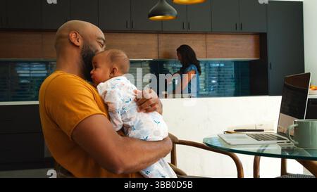Sorridente padre Cuddling e giocare con Baby Daughter a casa con madre in sottofondo Foto Stock