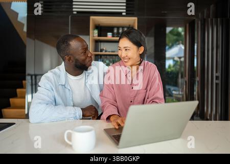 Felice coppia multietnica che utilizza un notebook, acquista online con le vendite, gioca insieme Foto Stock