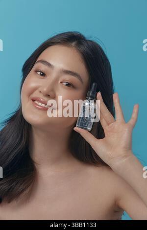 allegra donna di etnia giapponese che tiene il siero facciale vicino a sé stesso, esprime piacere e piacere potendo su uno sfondo blu, primi piani vertiali Foto Stock