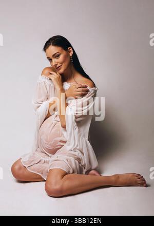 Ritratto di una bella donna incinta con capelli bagnati e tunica bianca bagnata che posa in studio su sfondo bianco Foto Stock