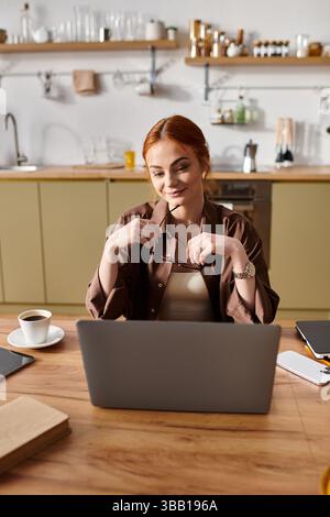 Una bellissima donna dai capelli rossi sorride mentre lavora al suo laptop in un ambiente accogliente in cucina. Foto Stock