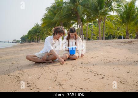Giovane coppia asiatica che si gode un momento di pace sulla spiaggia mentre si siede sulla sabbia e gioca con un secchio blu, catturando un senso di calma, closen Foto Stock