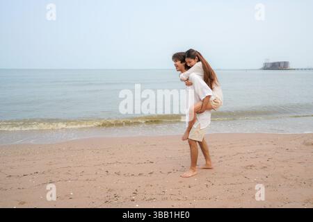 Giovane coppia asiatica che passa del tempo in una spiaggia tropicale, con l'uomo che fa un giro in piggyback alla donna lungo il litorale, esprimendo gioia, io Foto Stock