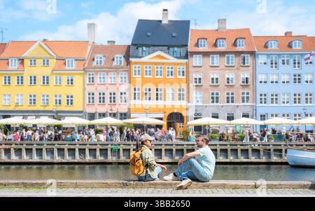 Coppia rilassati in riva all'acqua a Nyhavn, Copenaghen, Danimarca, prendendo il sole mentre edifici colorati riflettono il tranquillo fiume in una vivace città. L'atmosfera vivace cattura la gioia dell'estate. Foto Stock