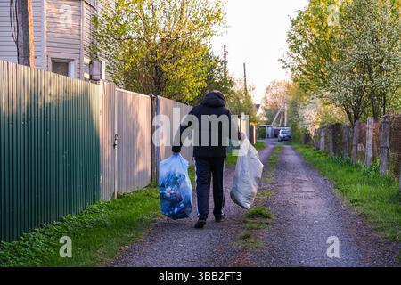 mosca, russia, 10.05.2025 Un uomo sta camminando lungo una strada con due sacchi di spazzatura. I sacchetti sono riempiti di bottiglie di plastica. Quell'uomo indossa una j nera Foto Stock